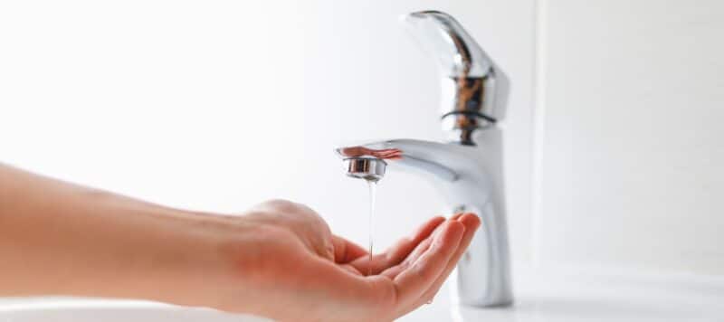 A close-up of a hand under a chrome faucet with a gentle stream of water flowing, illustrating water usage